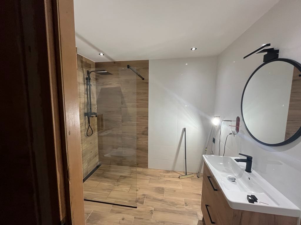 Nowoczesna łazienka z kabiną prysznicową typu walk-in, drewnopodobnymi płytkami i okrągłym lustrem z czarną ramą. Minimalistyczna armatura i oświetlenie punktowe.