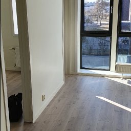 SERWIS DOMOWY - Jasne panele podłogowe w nowym mieszkaniu, w tle okno z widokiem na zewnątrz. W rogu wiertarka akumulatorowa i torba narzędziowa. Gniazdko elektryczne na ścianie.