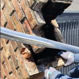 SERWIS DOMOWY - Demontaż uszkodzonego komina z cegły na rusztowaniu w Małdytach. Widoczne rękawice robocze i fragment samochodu na posesji. Prace murarskie.