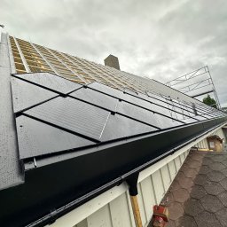 SERWIS DOMOWY - Montaż paneli fotowoltaicznych na dachu domu jednorodzinnego, widok z dołu na częściowo pokryty dach z widoczną konstrukcją nośną i rusztowaniem.