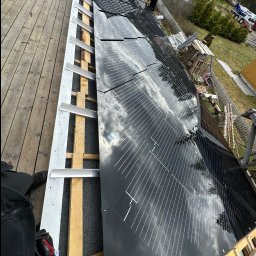 SERWIS DOMOWY - Montaż paneli słonecznych na dachu domu jednorodzinnego, widok z góry, częściowo widoczne rusztowanie i pracownik w oddali. Na panelach odbija się niebo.