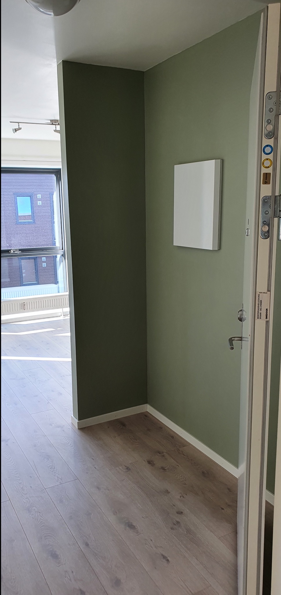 Wnętrze z zielonymi ścianami, jasną podłogą i białym panelem na ścianie obok drzwi. Widok na okno z grzejnikiem i budynkiem na zewnątrz.