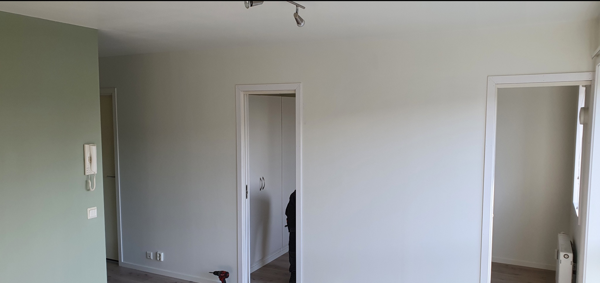 Jasne wnętrze po remoncie z widocznymi gniazdkami elektrycznymi, lampą sufitową i wiertarką przy ścianie. Świeżo pomalowane ściany i nowe drzwi.