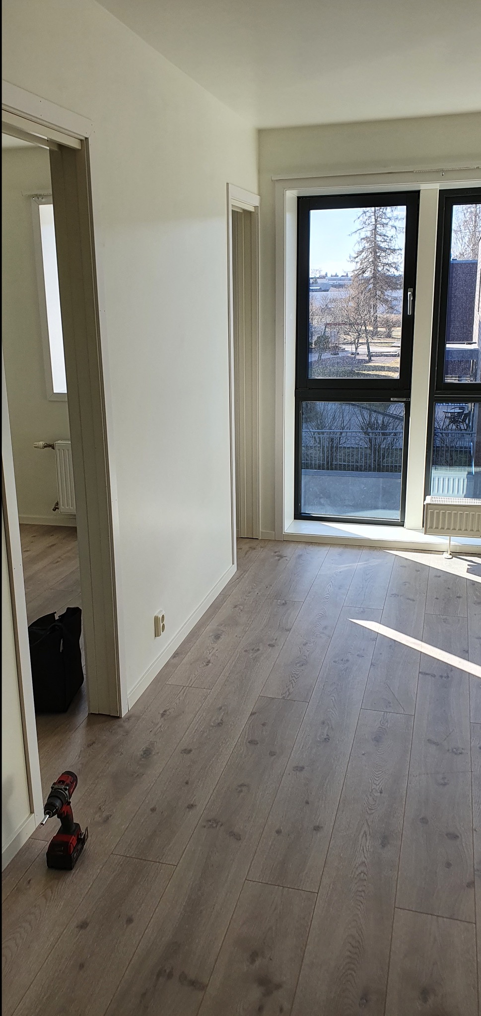 Jasne panele podłogowe w nowym mieszkaniu, w tle okno z widokiem na zewnątrz. W rogu wiertarka akumulatorowa i torba narzędziowa. Gniazdko elektryczne na ścianie.