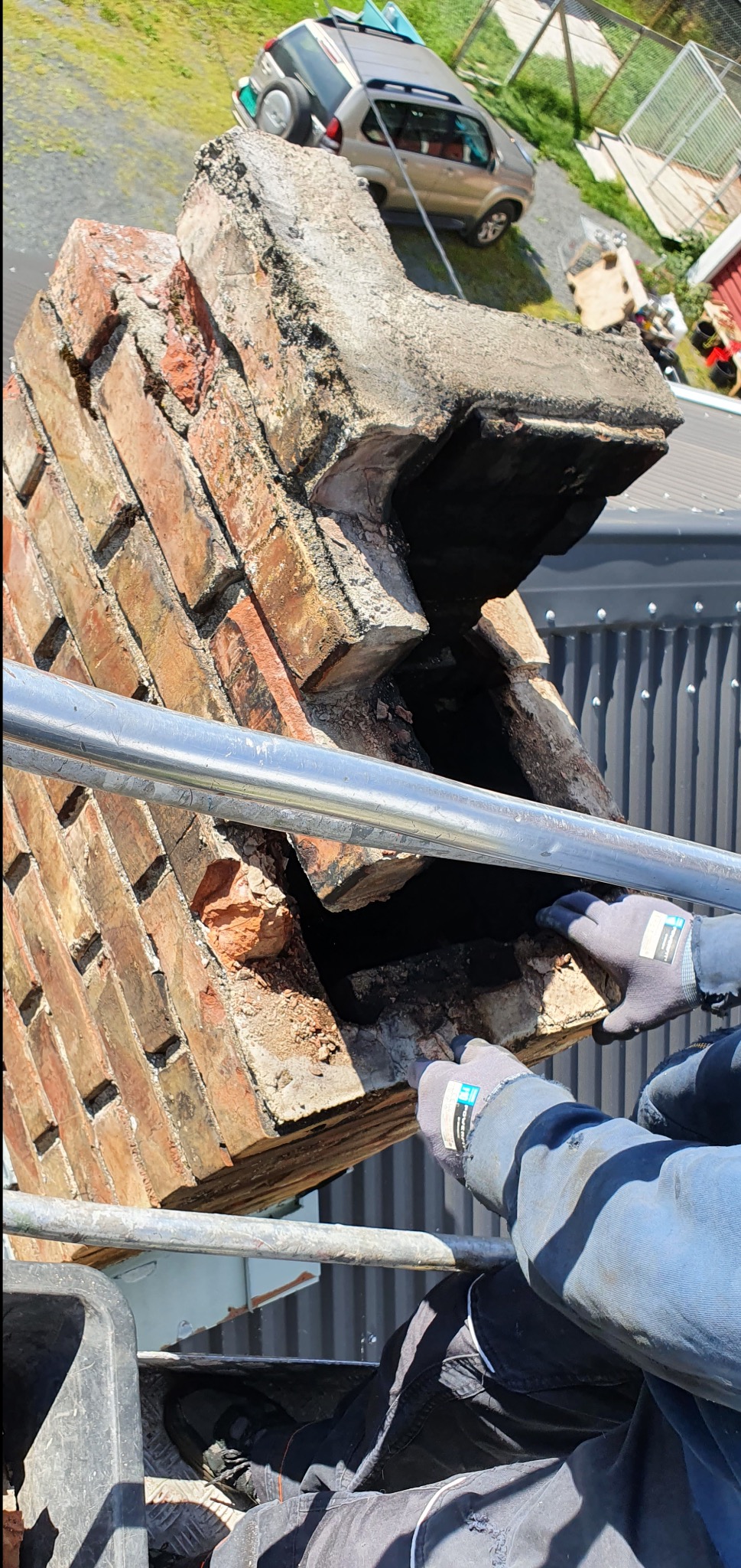 Demontaż uszkodzonego komina z cegły na rusztowaniu w Małdytach. Widoczne rękawice robocze i fragment samochodu na posesji. Prace murarskie.