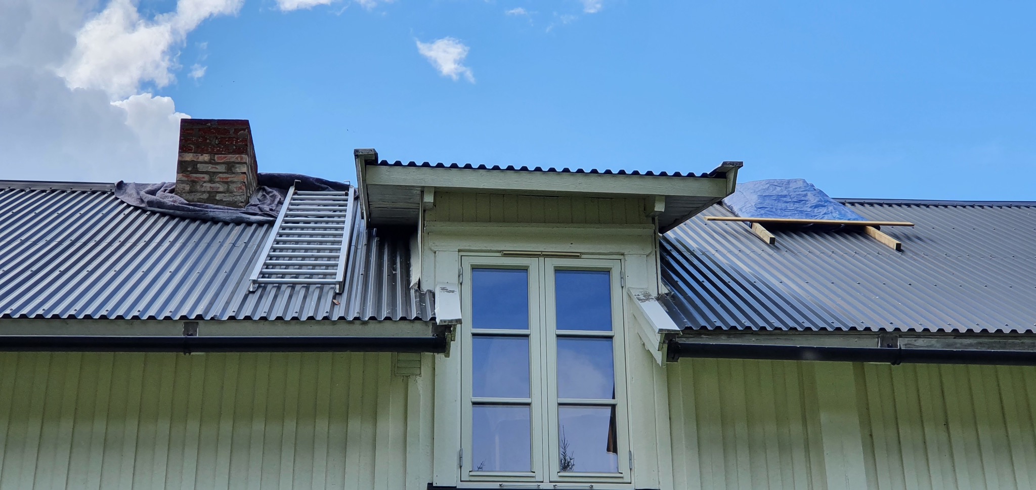 Falista blacha na dachu domu z oknem mansardowym, drabina oparta o komin. Fragment dachu zabezpieczony plandeką. Prace dekarskie w toku, widok z dołu.