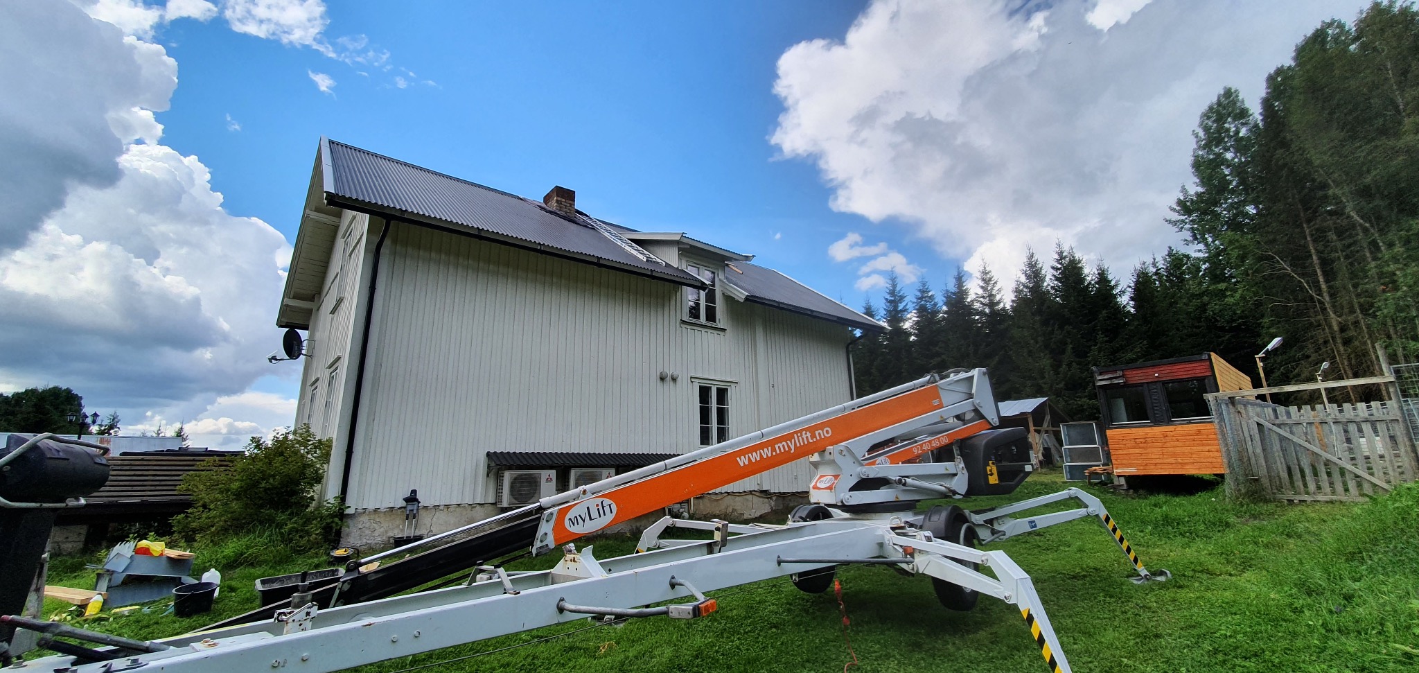 Biały dom z ciemnym dachem widziany z dołu, z podnośnikiem koszowym MyLift na pierwszym planie. Widoczne okna, klimatyzatory i fragment lasu w tle.