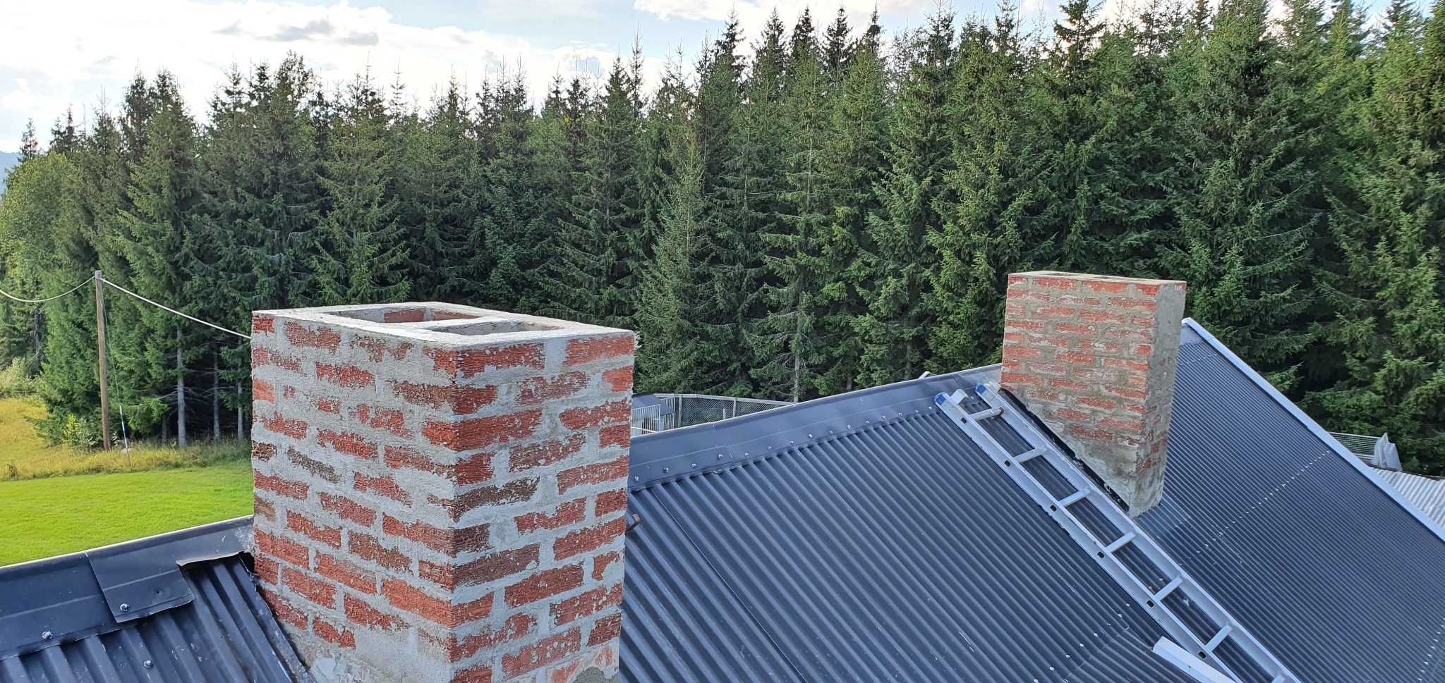 Ceglane kominy na metalowym dachu z drabiną, na tle zielonego lasu. Jeden komin z pustką w środku, drugi mniejszy. Widok z góry, w słoneczny dzień.