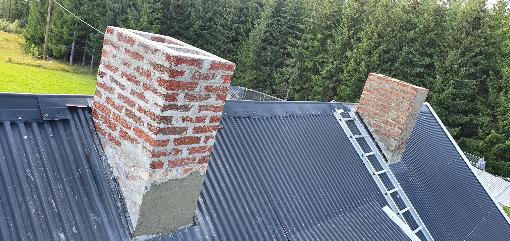 Odnowiony komin z cegły na dachu z blachy trapezowej, z drabinką i zielonym tłem drzew. Widoczne ślady naprawy i świeża zaprawa.
