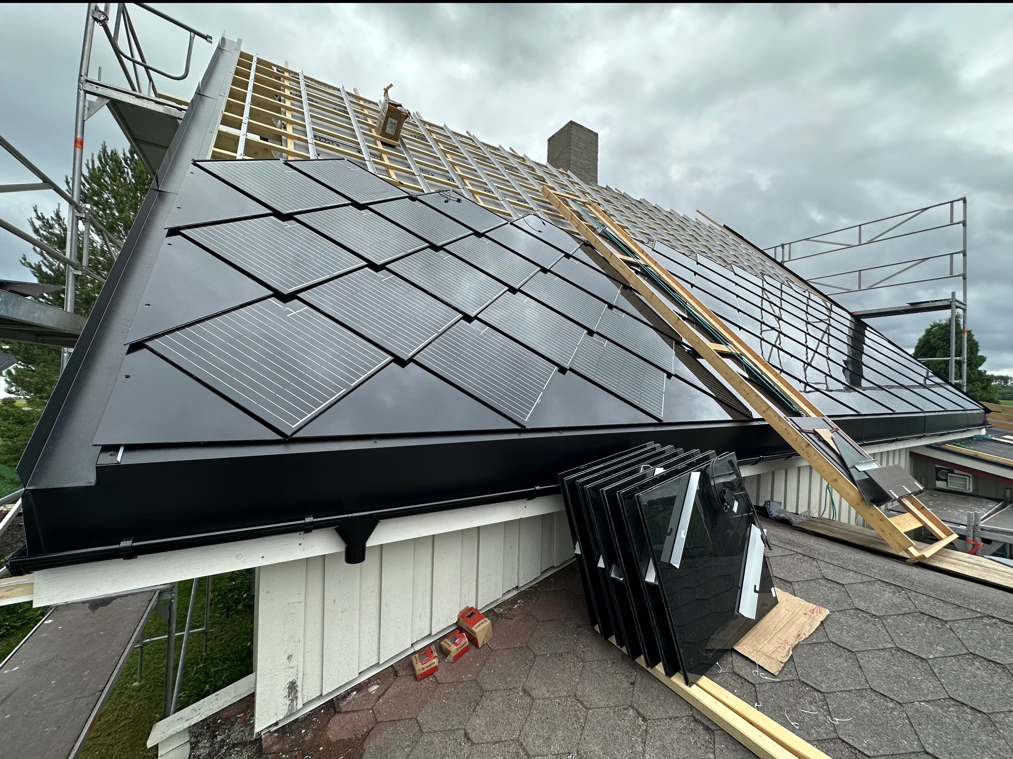 Montaż paneli fotowoltaicznych na dachu domu z użyciem rusztowania. Część paneli już zamontowana, pozostałe czekają na ułożenie. Widoczne elementy konstrukcji dachu.