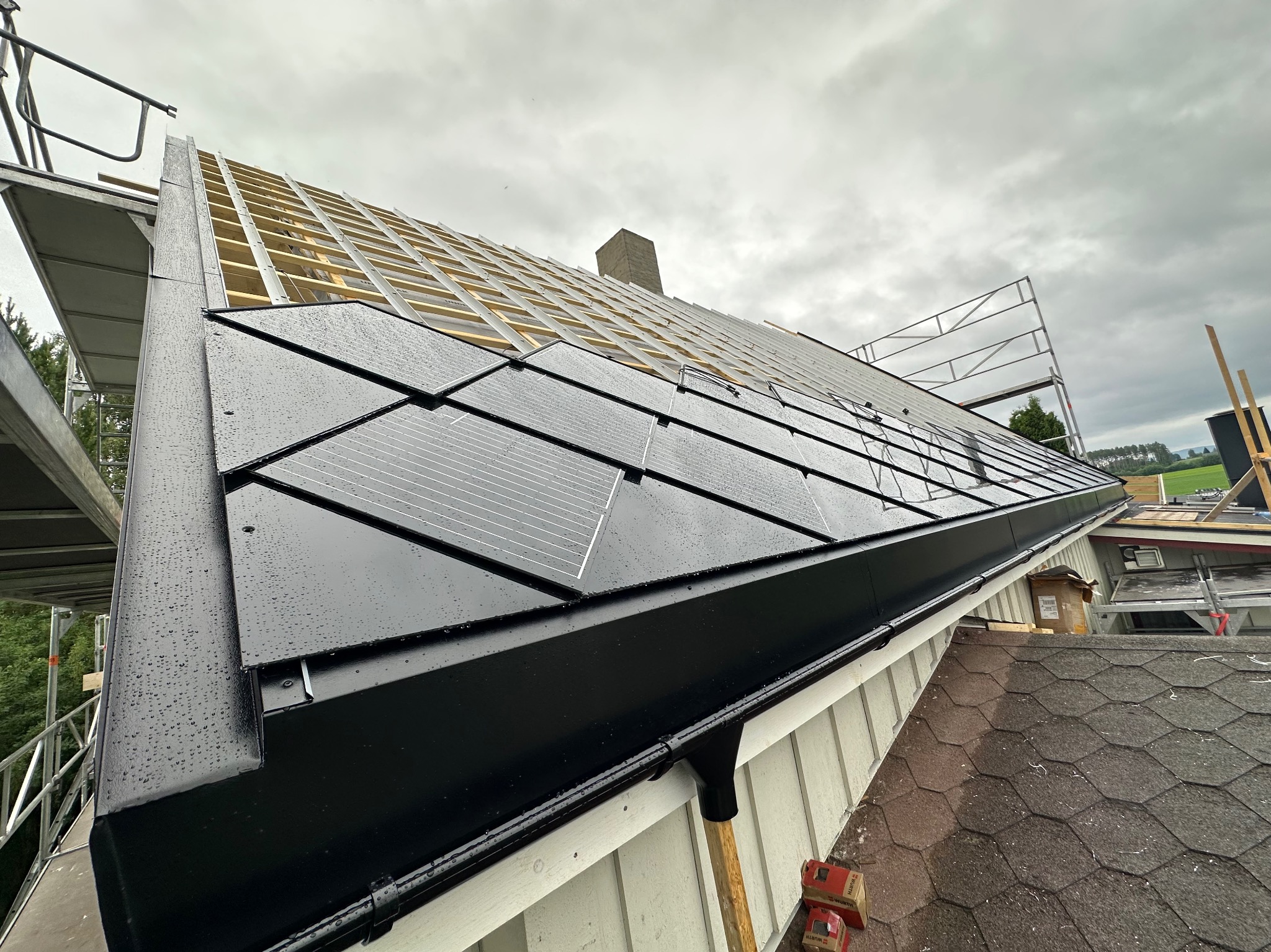 Montaż paneli fotowoltaicznych na dachu domu jednorodzinnego, widok z dołu na częściowo pokryty dach z widoczną konstrukcją nośną i rusztowaniem.