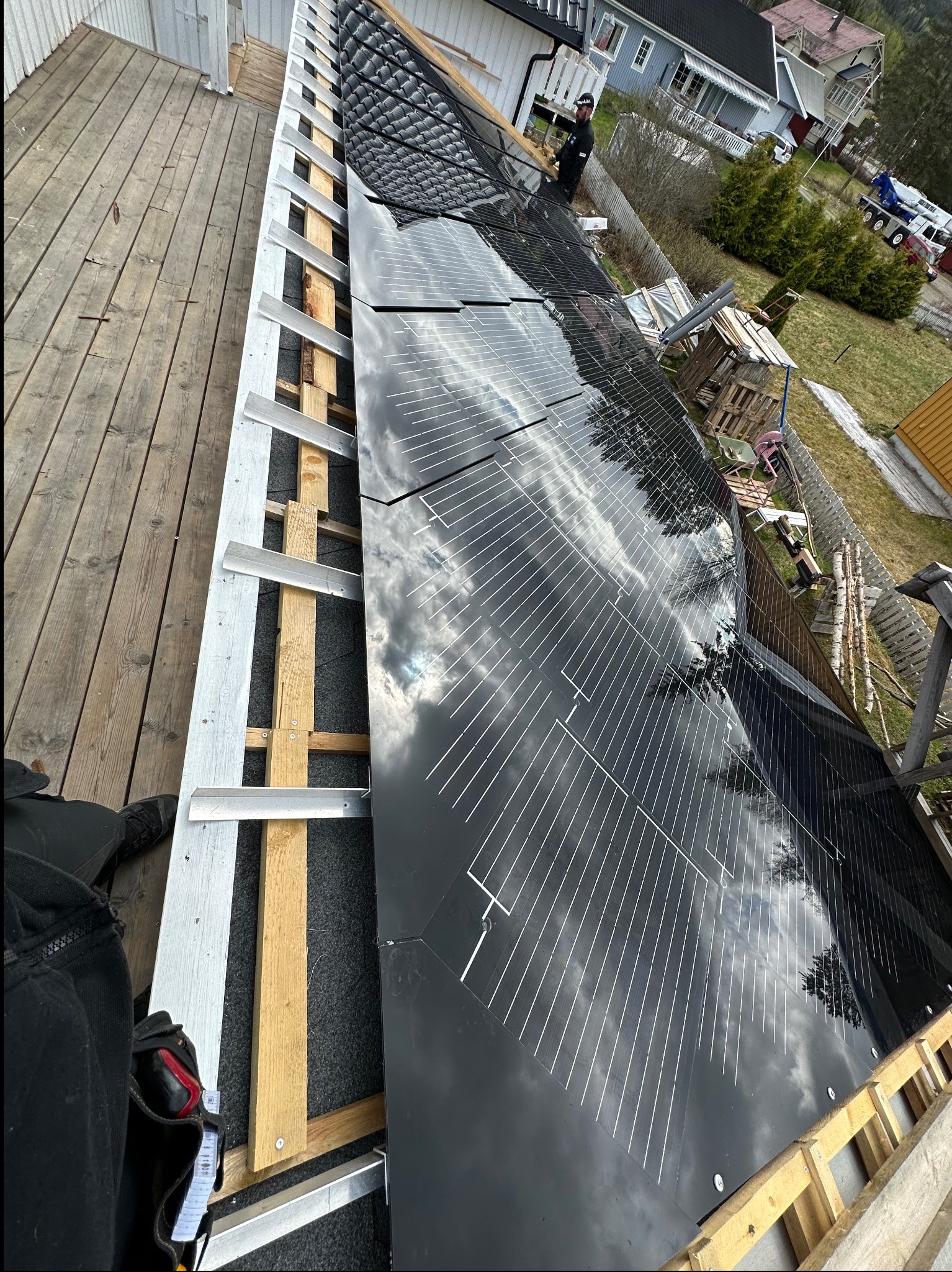 Montaż paneli słonecznych na dachu domu jednorodzinnego, widok z góry, częściowo widoczne rusztowanie i pracownik w oddali. Na panelach odbija się niebo.
