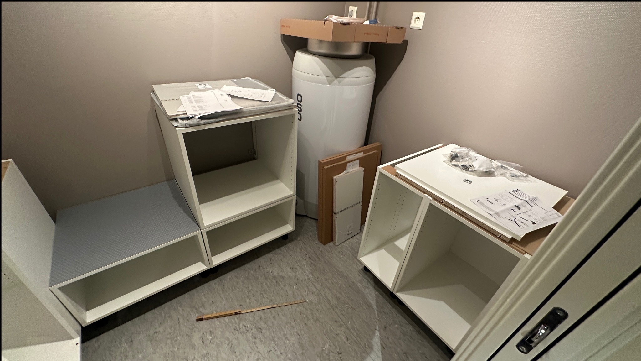 Rozpakowane, białe szafki kuchenne z instrukcjami i elementami montażowymi, gotowe do instalacji w pomieszczeniu z bojlerem. Widoczny fragment miarki.