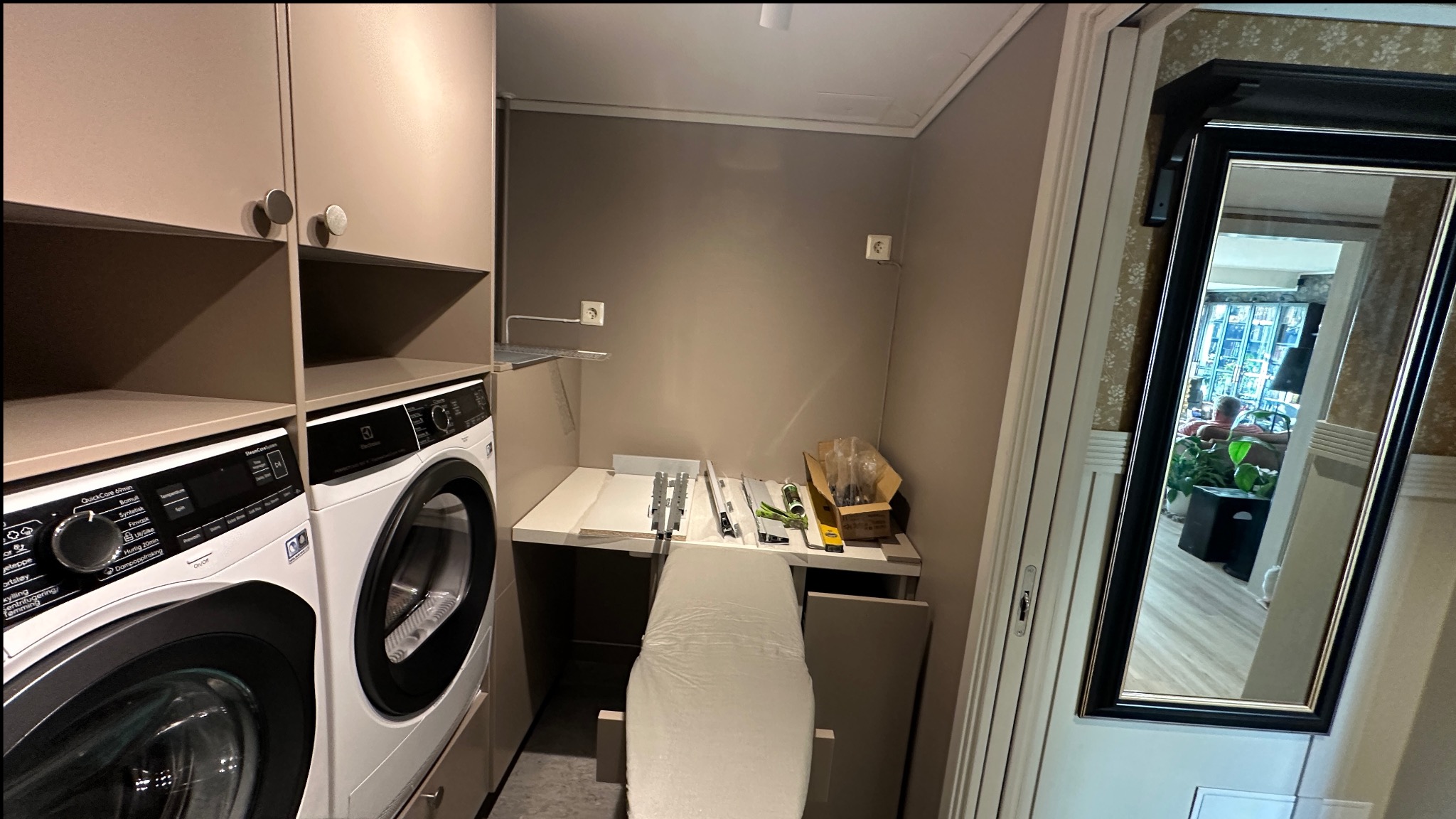 Pralka i suszarka w zabudowie meblowej, obok rozłożona deska do prasowania i elementy do montażu mebli. Widoczne gniazdka elektryczne na ścianie.