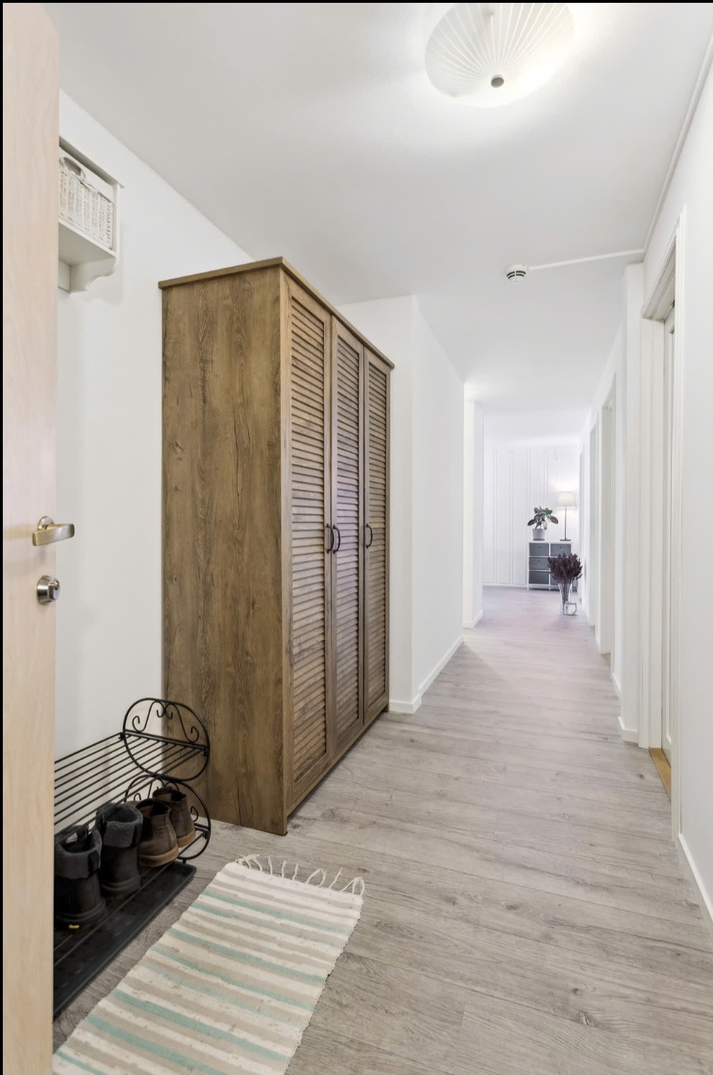 Jasny korytarz z drewnianą szafą z żaluzjowymi drzwiami, metalową półką na buty i dywanikiem w paski. Na suficie widoczny czujnik dymu i lampa.