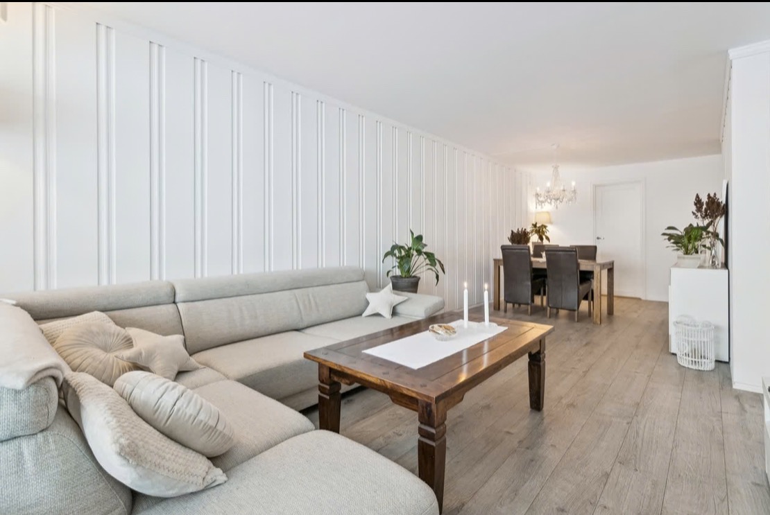Jasne wnętrze salonu z drewnianym stołem, szarą sofą i dekoracyjną ścianą z białych paneli. Widoczny parkiet i elementy oświetlenia.