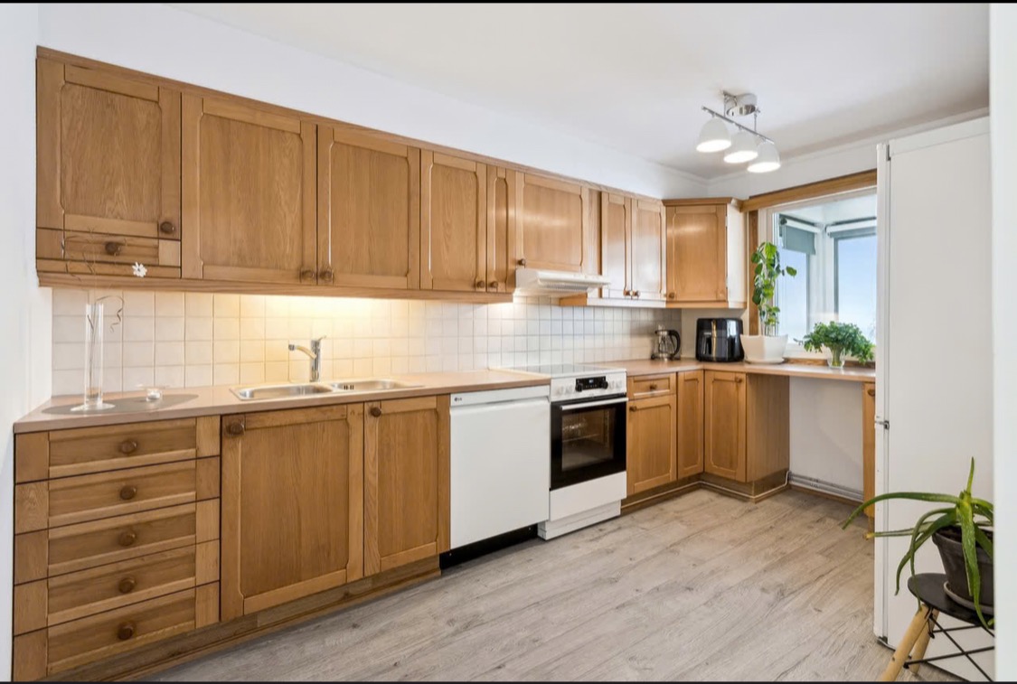 Jasna kuchnia z drewnianymi szafkami, białymi płytkami i sprzętami. Widoczne okno z roślinami i szare panele podłogowe. Oświetlenie sufitowe z trzema lampami.