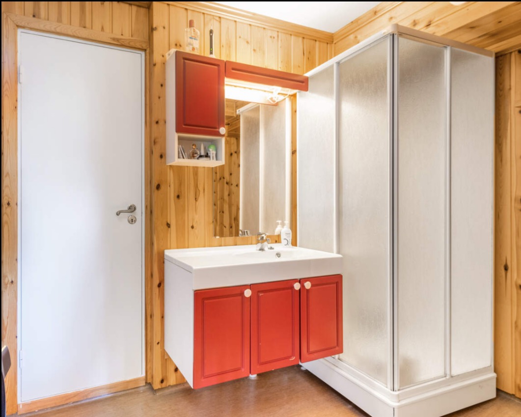 Jasna łazienka z drewnianą boazerią, kabiną prysznicową i umywalką z czerwoną szafką. Umywalka oświetlona lampą. Widok na białe drzwi.