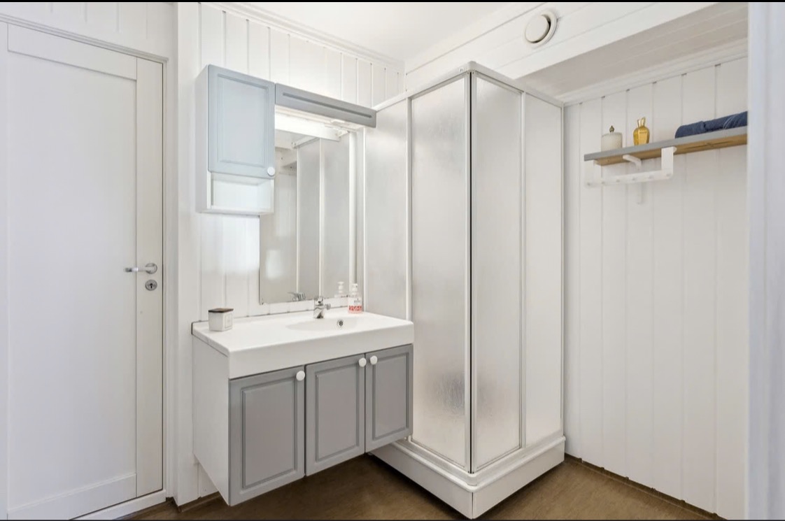 Jasna łazienka z kabiną prysznicową i umywalką z szafką w odcieniach szarości. Białe panele ścienne i drzwi. Minimalistyczny design z drewnianą podłogą.