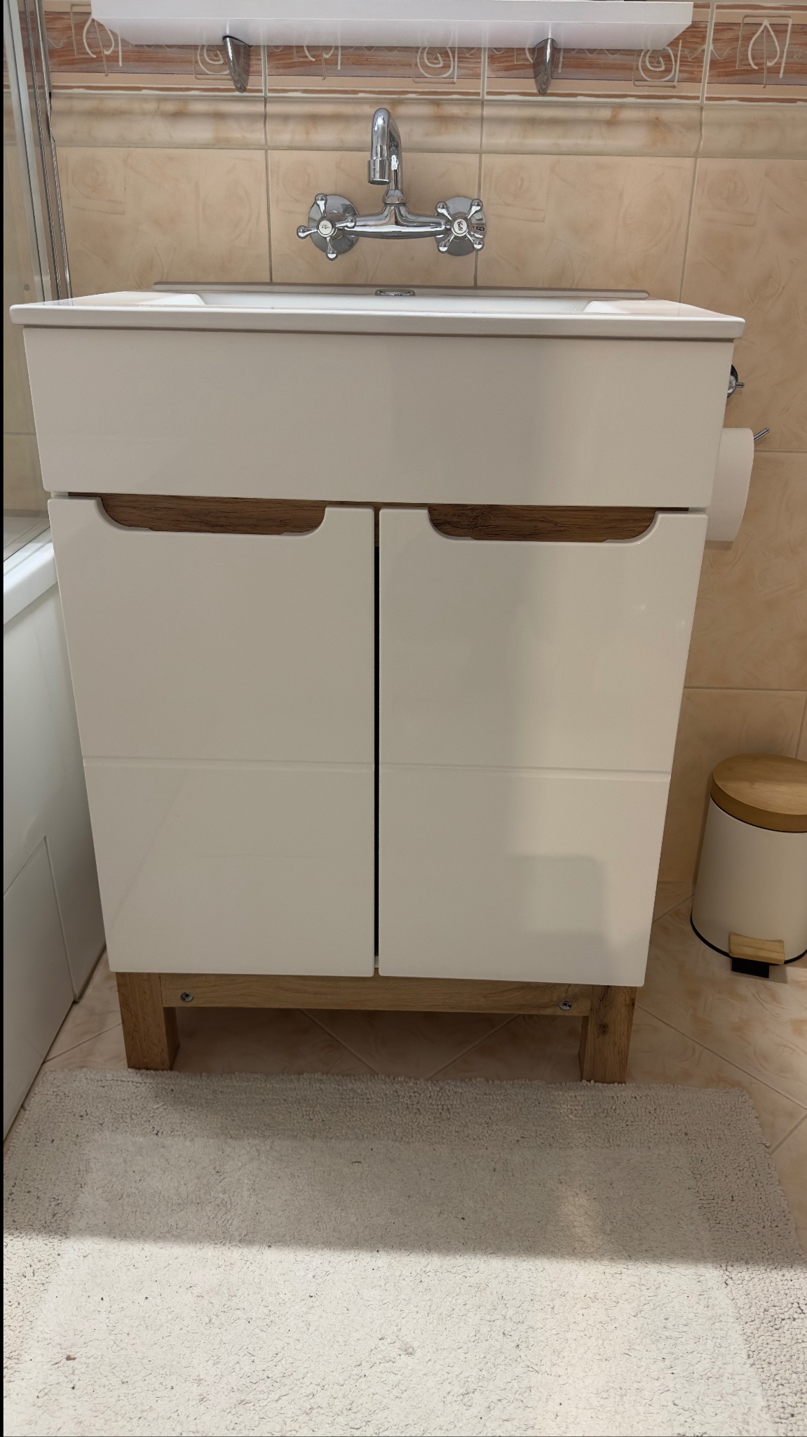 Biała szafka pod umywalkę z drewnianymi akcentami i chromowaną baterią na tle beżowych płytek. Widoczny dywanik i kosz na śmieci.