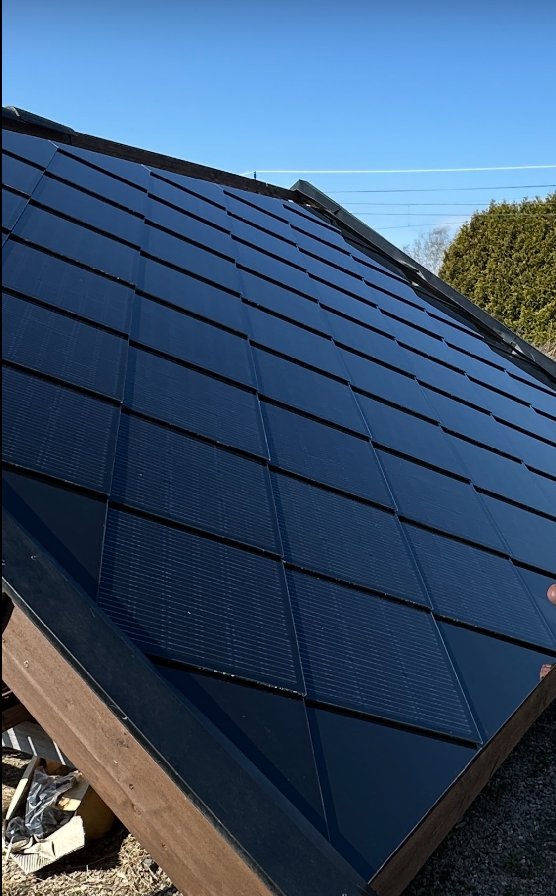Błyszczące, czarne panele fotowoltaiczne w kształcie dachówek, zamontowane na skośnym dachu w słoneczny dzień. Widoczne elementy konstrukcyjne dachu i fragment zieleni w tle.