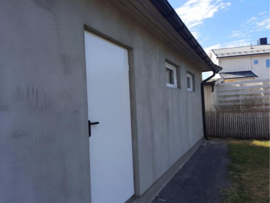 Szara ściana budynku z białą stolarką okienną i drzwiami, czarną rynną i fragmentem ogrodzenia w tle, w słoneczny dzień. Widok z zewnątrz.