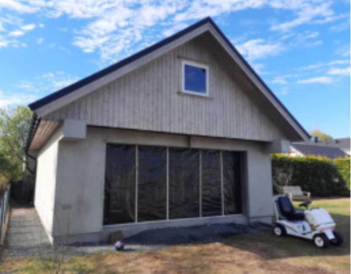 Garaż z drewnianą elewacją i oknem, zabezpieczony folią. Widoczny trawnik, niebieskie niebo i wózek inwalidzki. Prace wykończeniowe w słoneczny dzień.