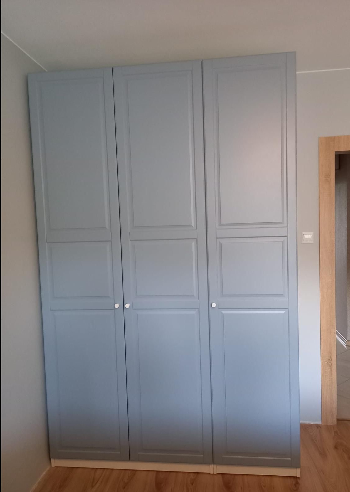 Trzydrzwiowa, szara szafa z panelowymi drzwiami i białymi gałkami, ustawiona na drewnianej podłodze w jasnym pokoju. Widoczna rama drzwi po prawej.