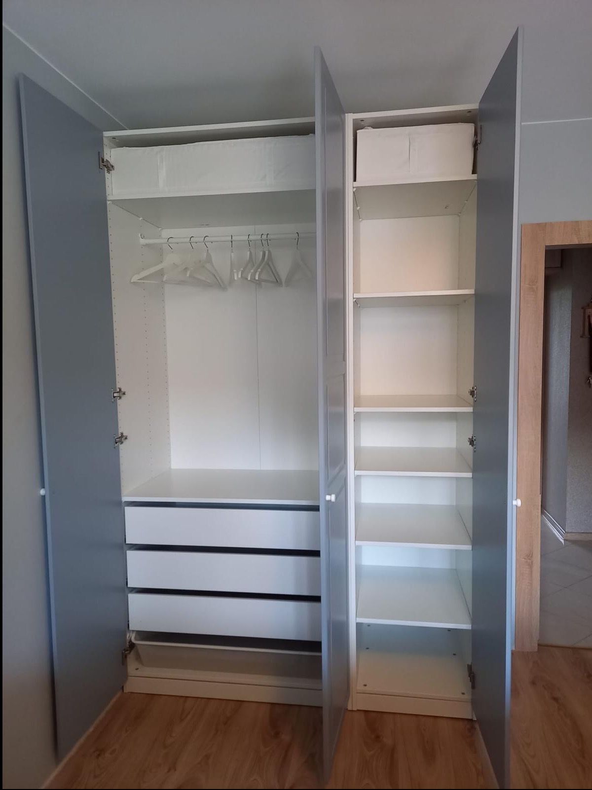 Wnętrze szafy wnękowej z szufladami, półkami i miejscem na wieszaki. Drzwi szafy w kolorze szarym, wnętrze białe. Widoczna podłoga z paneli.