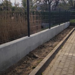 FIRMA HANDLOWA RO - Producent siatki ogrodzeniowej - Nowoczesne ogrodzenie panelowe z podmurówką betonową, widok z boku, zarośla w tle. Elementy metalowe w kolorze grafitowym, kostka brukowa obok. Słoneczny dzień.
