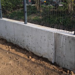 FIRMA HANDLOWA RO - Producent siatki ogrodzeniowej - Nowoczesne ogrodzenie panelowe z podmurówką betonową, widok z bliska na świeżo obsypaną ziemią podstawę. W tle zieleń i zaparkowane pojazdy.