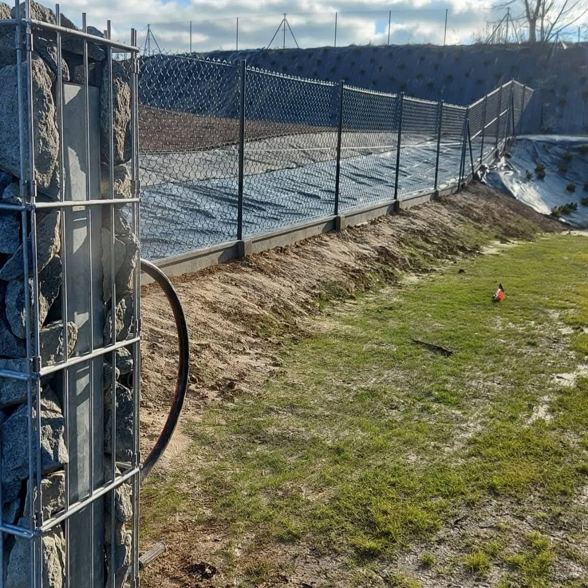 Nowoczesne ogrodzenie z siatki i gabionów na betonowej podmurówce, zabezpieczające teren. Widok na wzgórze w tle, słoneczny dzień.