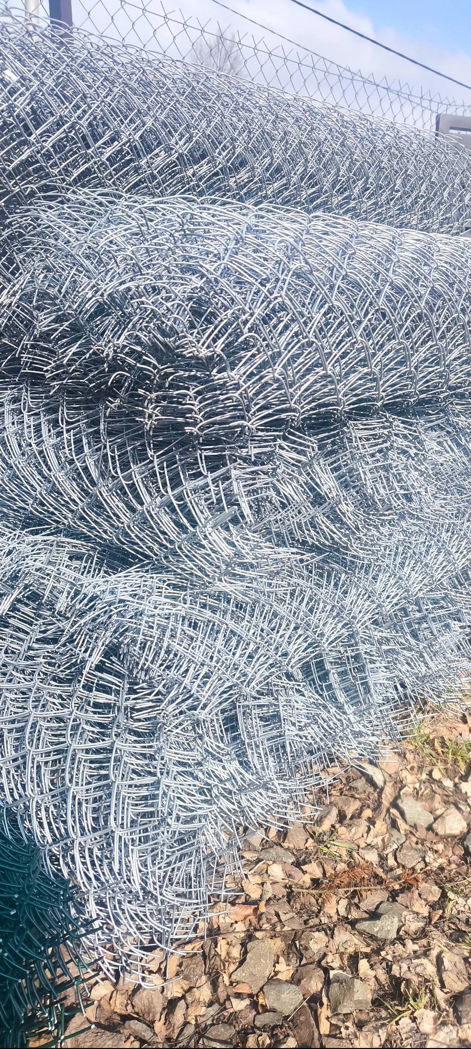 Stos rolek srebrnej siatki ogrodzeniowej na ziemi z kamieniami i liśćmi. Widok z bliska, ukazujący strukturę i detale materiału.