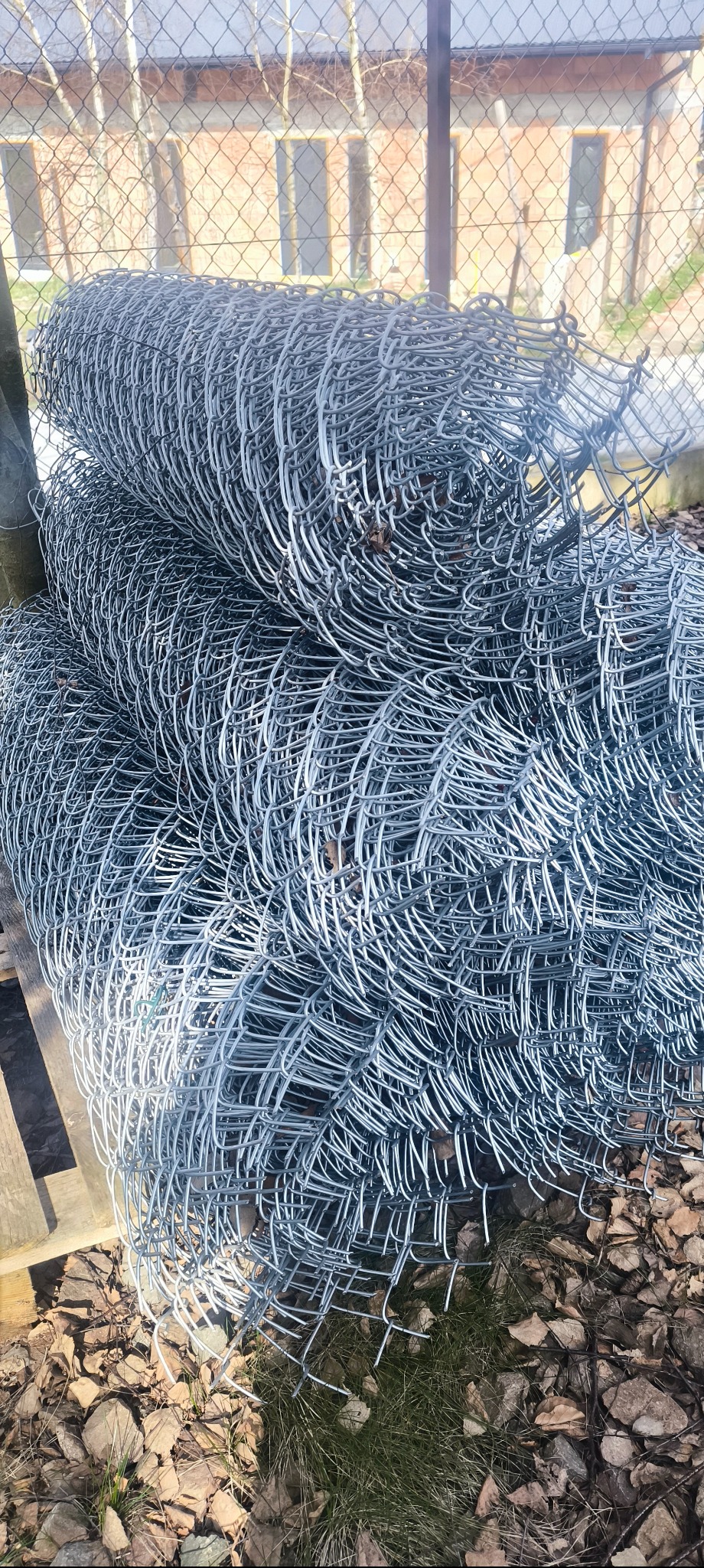 Stos rolek srebrnej siatki ogrodzeniowej na palecie, z widocznym tłem budynku z cegły przez oczka siatki. Drobne liście i trawa u podstawy palety.