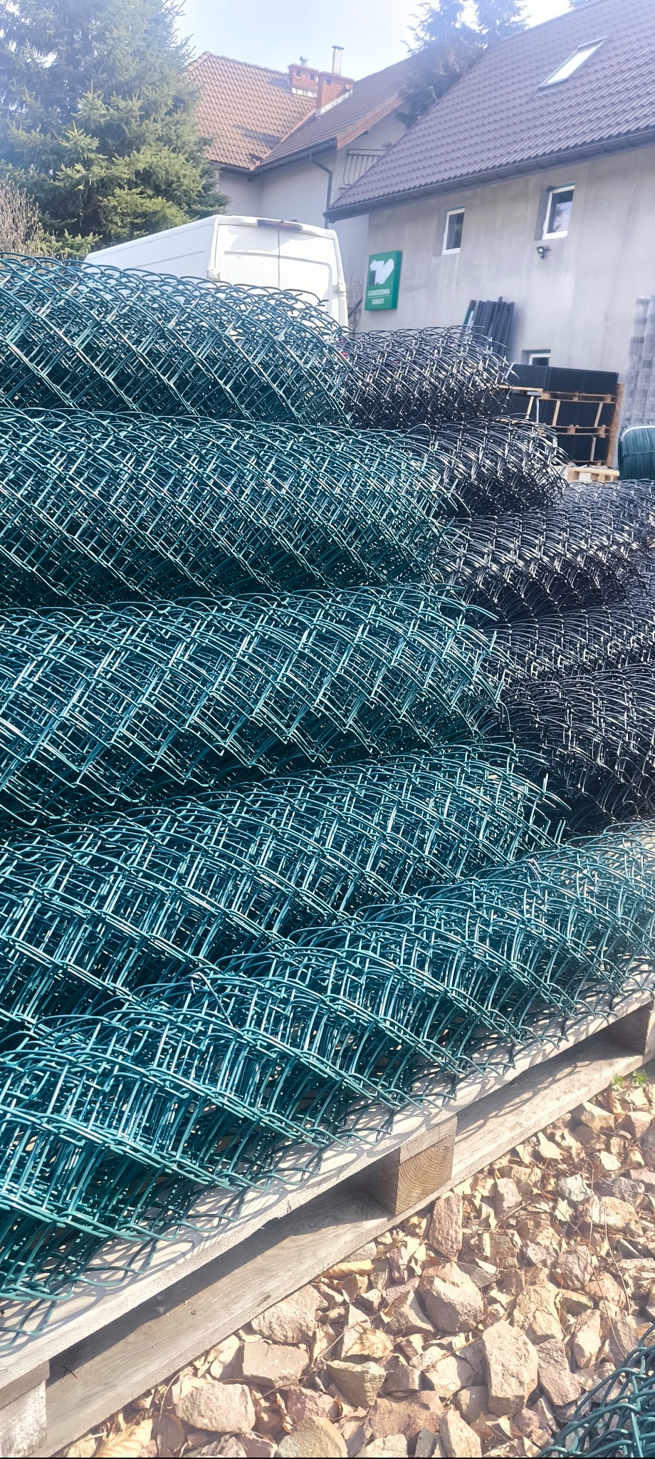 Zestaw zwiniętej siatki ogrodzeniowej w kolorze zielonym i grafitowym, ułożonej na drewnianej palecie. W tle budynek z reklamą.