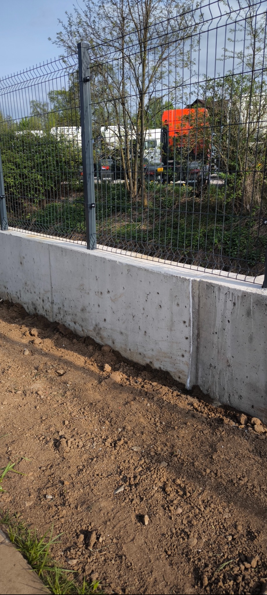 Nowoczesne ogrodzenie panelowe z podmurówką betonową, widok z bliska na świeżo obsypaną ziemią podstawę. W tle zieleń i zaparkowane pojazdy.
