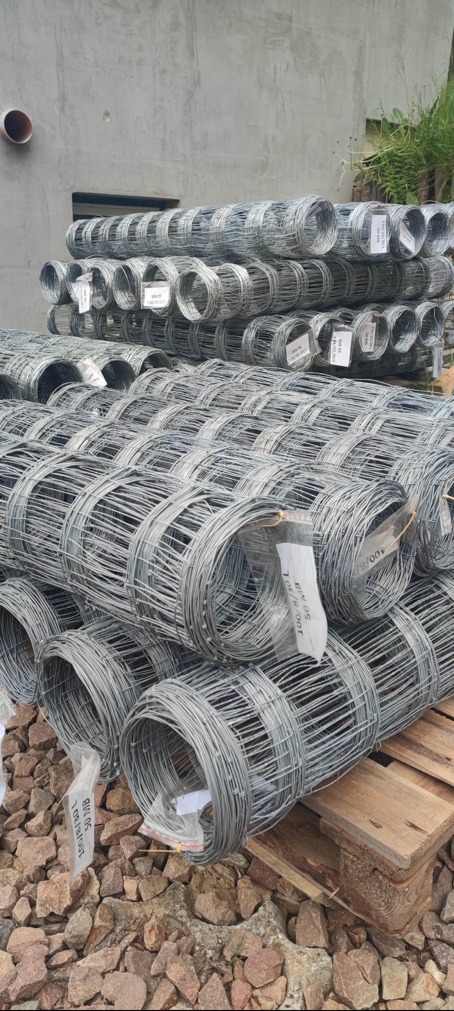 Stos rolek siatki ogrodzeniowej na palecie, z etykietami informacyjnymi. Widoczne kamienie i betonowe tło. Ujęcie z bliska, ukazujące strukturę siatki.