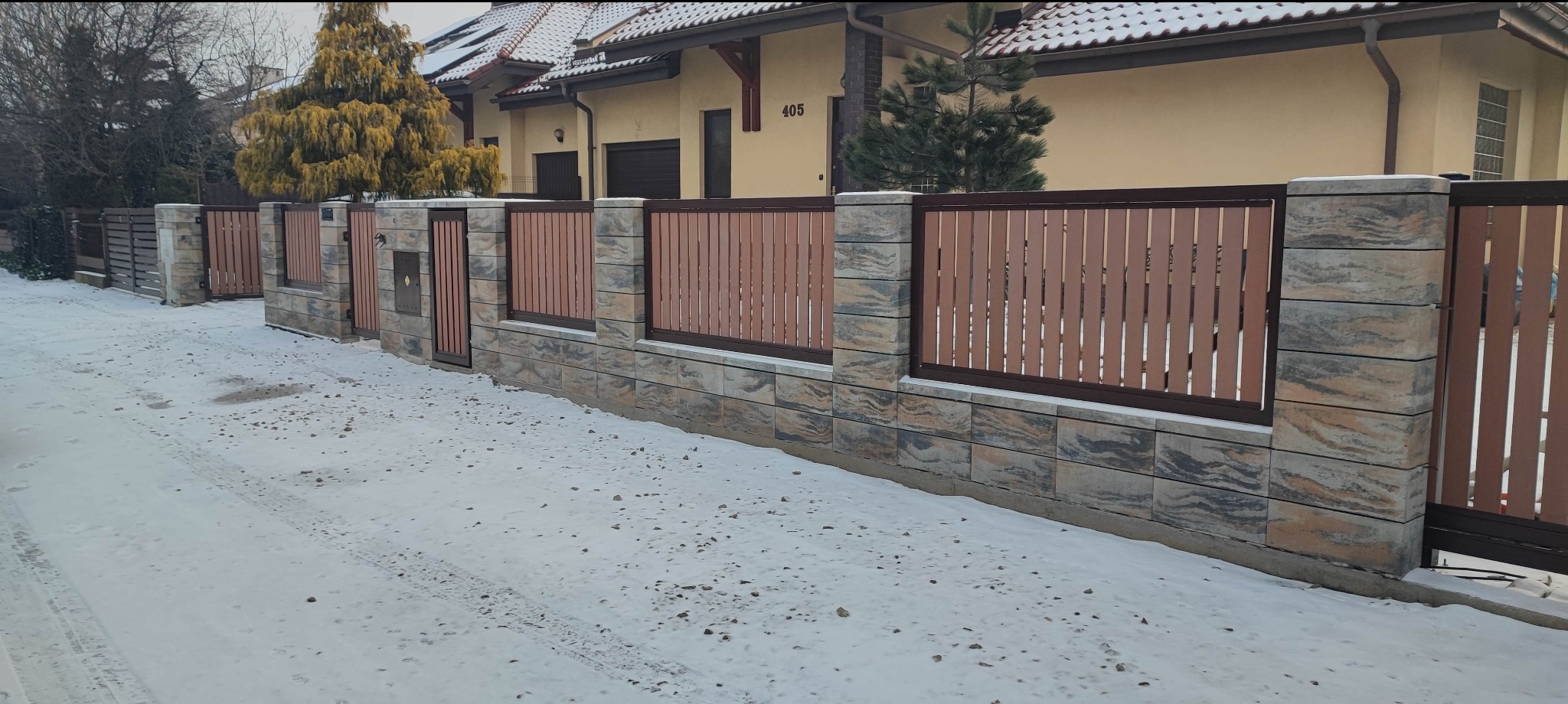 Nowoczesne ogrodzenie z elementami betonowymi i drewnianymi lamelami w brązowym odcieniu, brama wejściowa, zimowy krajobraz z ośnieżonym terenem.