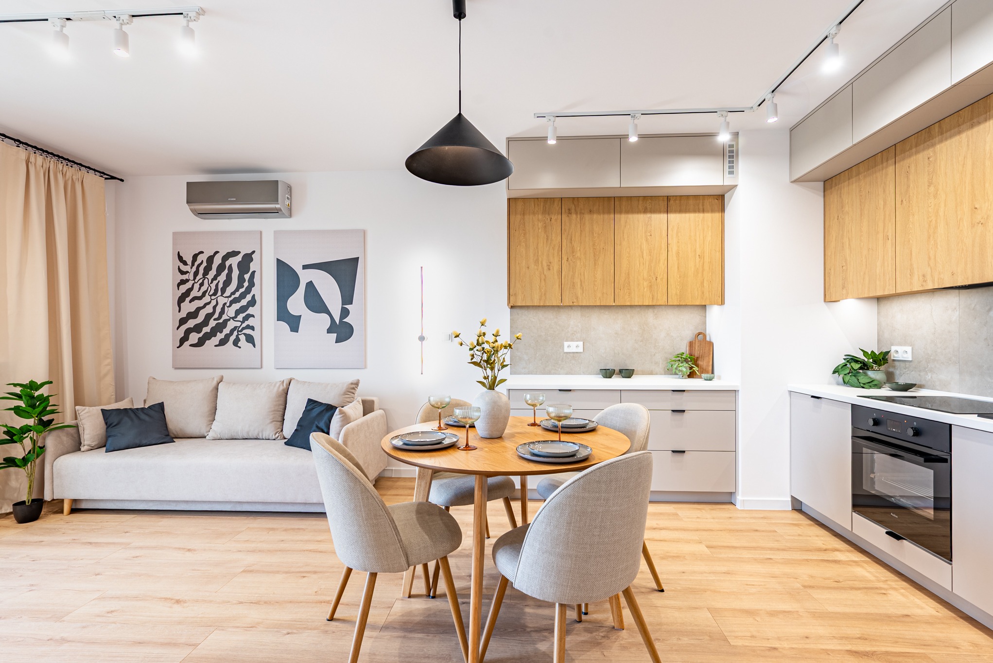 Jasne, nowocześnie urządzone wnętrze: salon z aneksem kuchennym. Okrągły stół z krzesłami, sofa z poduszkami, minimalistyczne obrazy na ścianie i drewniana podłoga.