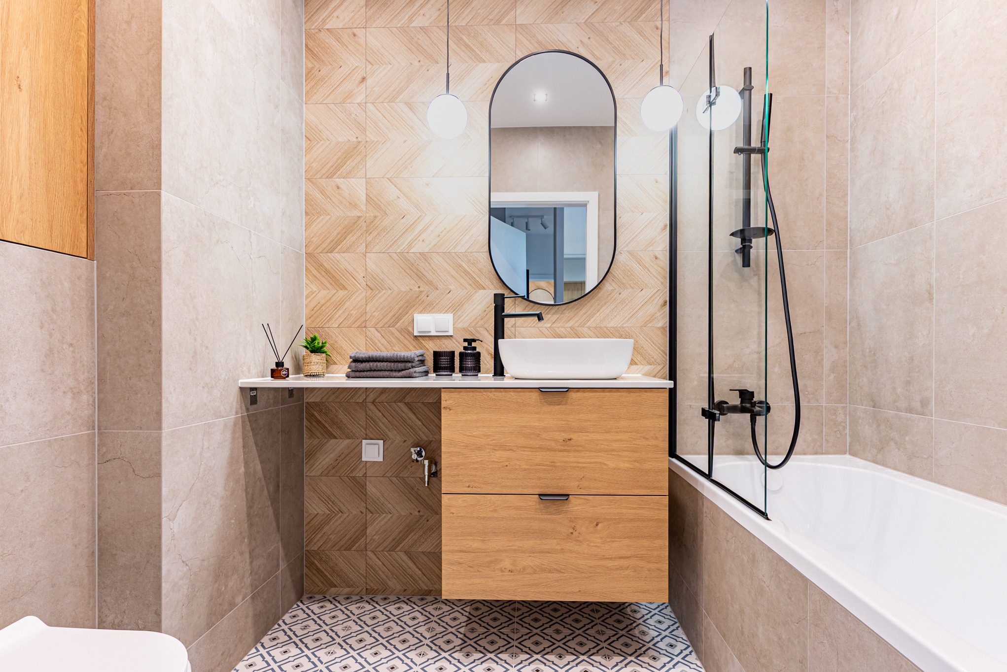 Nowoczesna łazienka z drewnianymi akcentami: szafka pod umywalką, mozaika na podłodze i geometryczny wzór na ścianie. Minimalistyczny design z czarną armaturą.