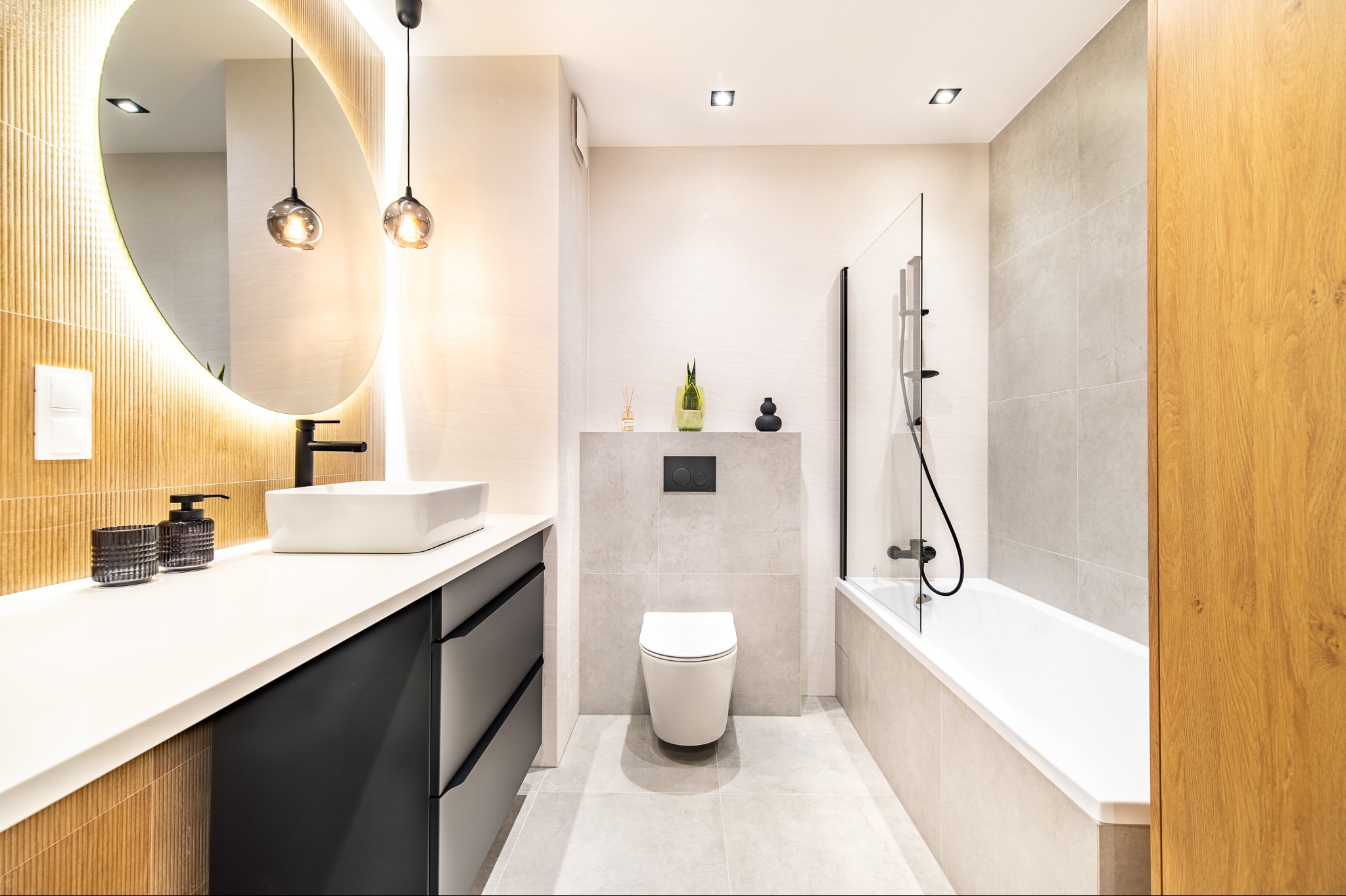 Nowoczesna łazienka z owalnym lustrem podświetlanym LED, białą umywalką nablatową i czarną armaturą. Jasne płytki i drewniane akcenty tworzą elegancki wystrój.