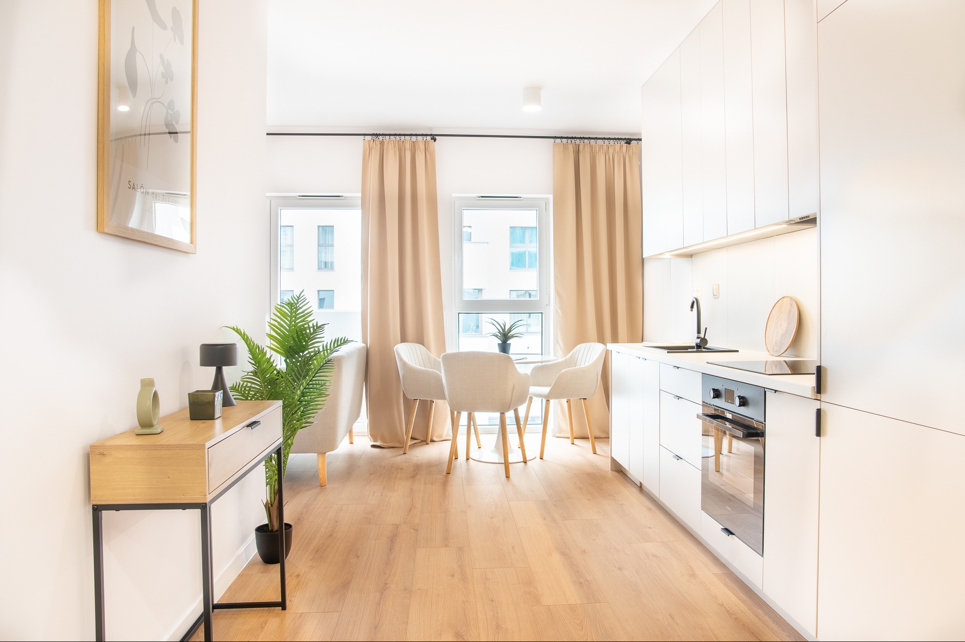 Jasne wnętrze: minimalistyczna kuchnia z drewnianą podłogą, białymi meblami, stołem z krzesłami i dekoracyjną konsolą z rośliną. Okna z beżowymi zasłonami.