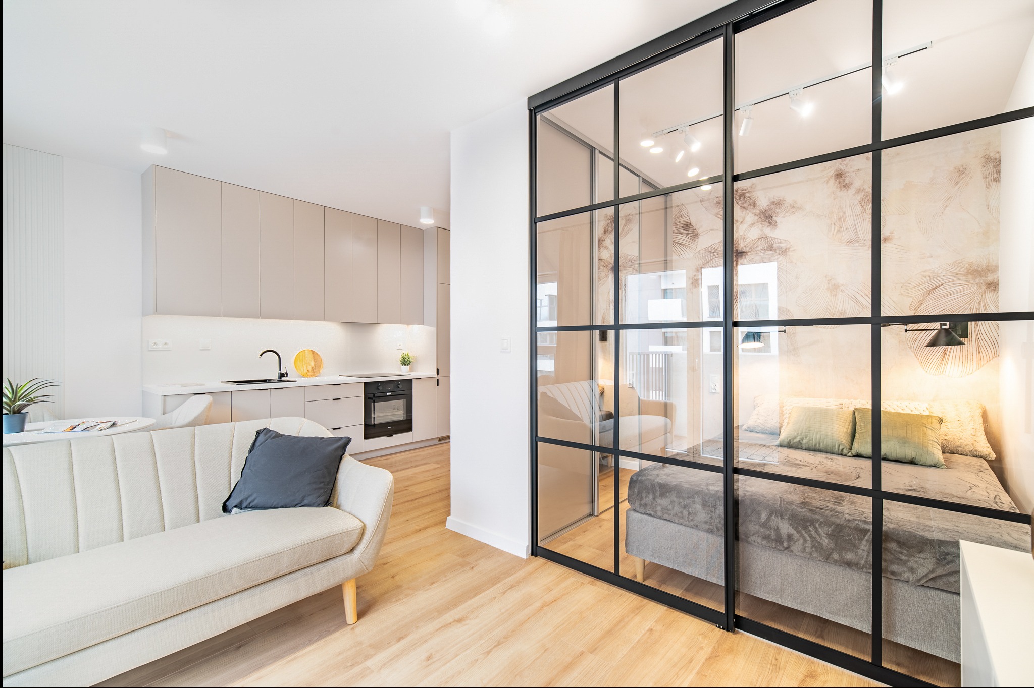 Jasne, nowoczesne studio z przeszkloną ścianą działową oddzielającą sypialnię od salonu z aneksem kuchennym. Minimalistyczny design, drewniana podłoga, sofa w odcieniach beżu.