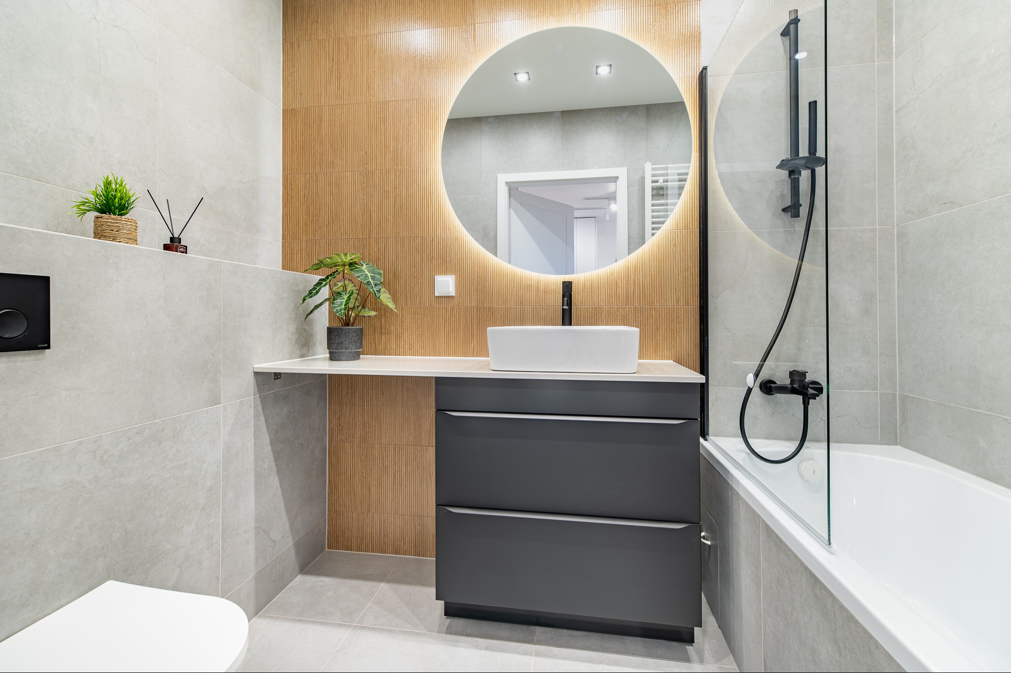 Nowoczesna łazienka z szarymi płytkami, drewnopodobną ścianą, okrągłym lustrem z podświetleniem LED, białą umywalką nablatową i czarną armaturą.