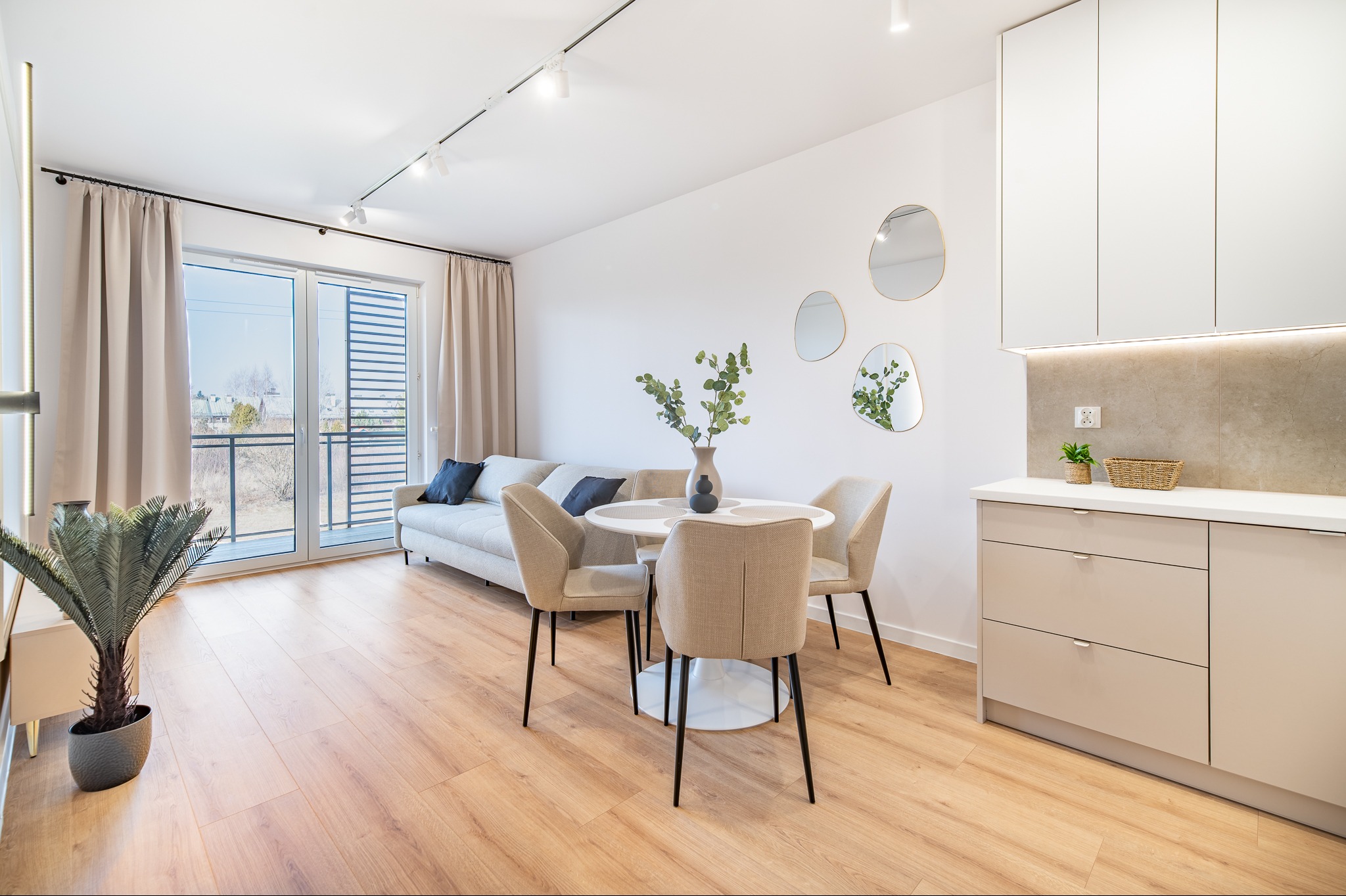 Jasne wnętrze: salon z aneksem kuchennym, drewniana podłoga, sofa, okrągły stół z krzesłami, minimalistyczne lustra i oświetlenie punktowe. Nowoczesny design.