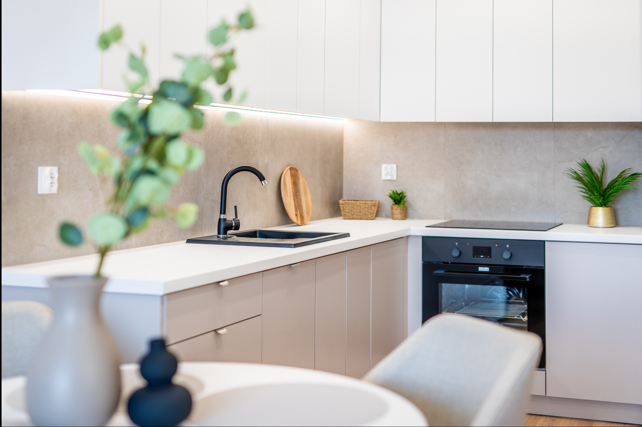 Nowoczesna kuchnia w jasnych barwach z czarną armaturą i drewnianymi akcentami. Minimalistyczny design, oświetlenie LED pod szafkami, roślina doniczkowa na blacie.