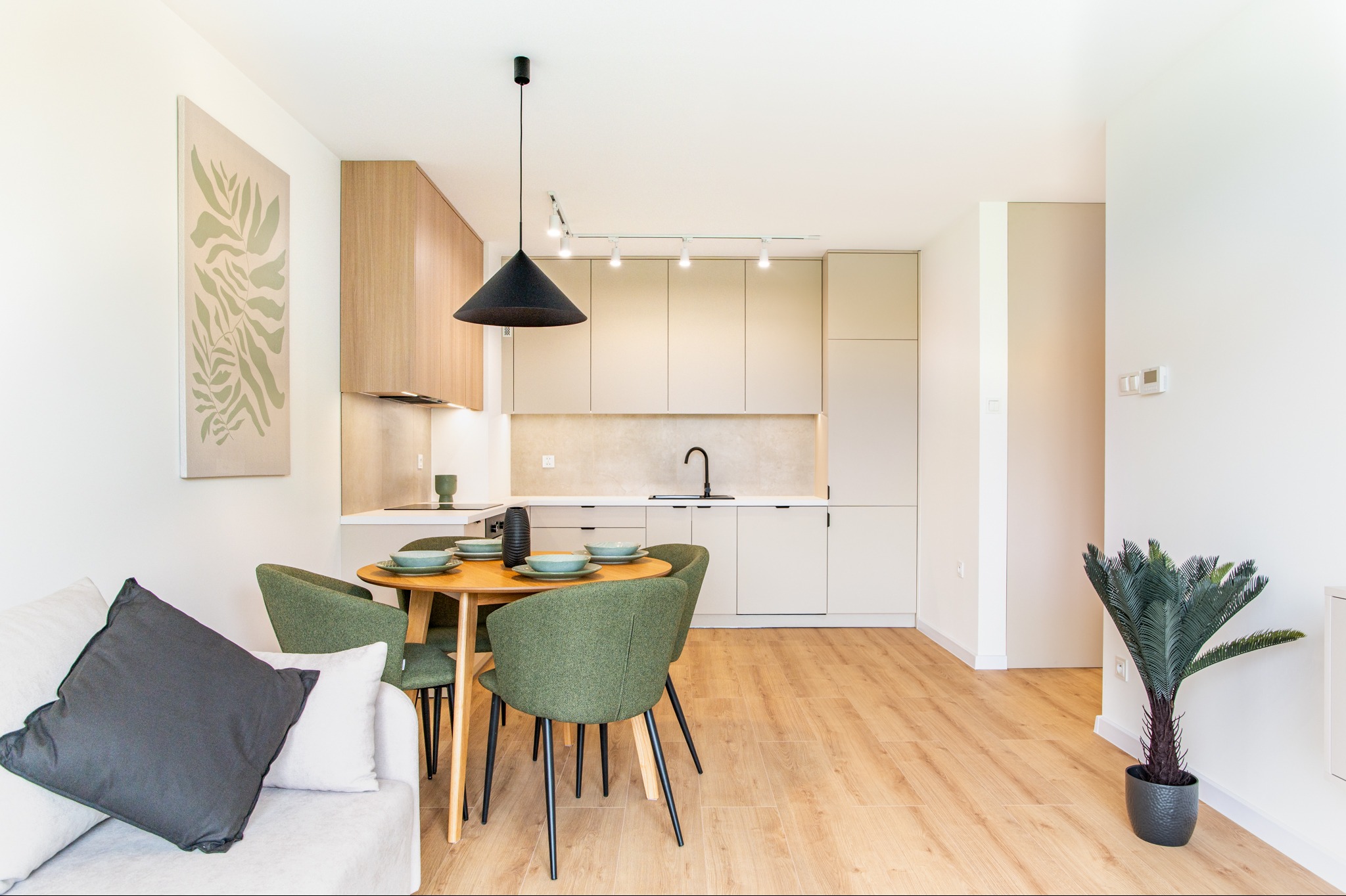 Jasne wnętrze: kuchnia z jadalnią w stylu skandynawskim. Drewniany stół z zielonymi krzesłami, beżowe szafki kuchenne, drewniana podłoga i roślina doniczkowa.