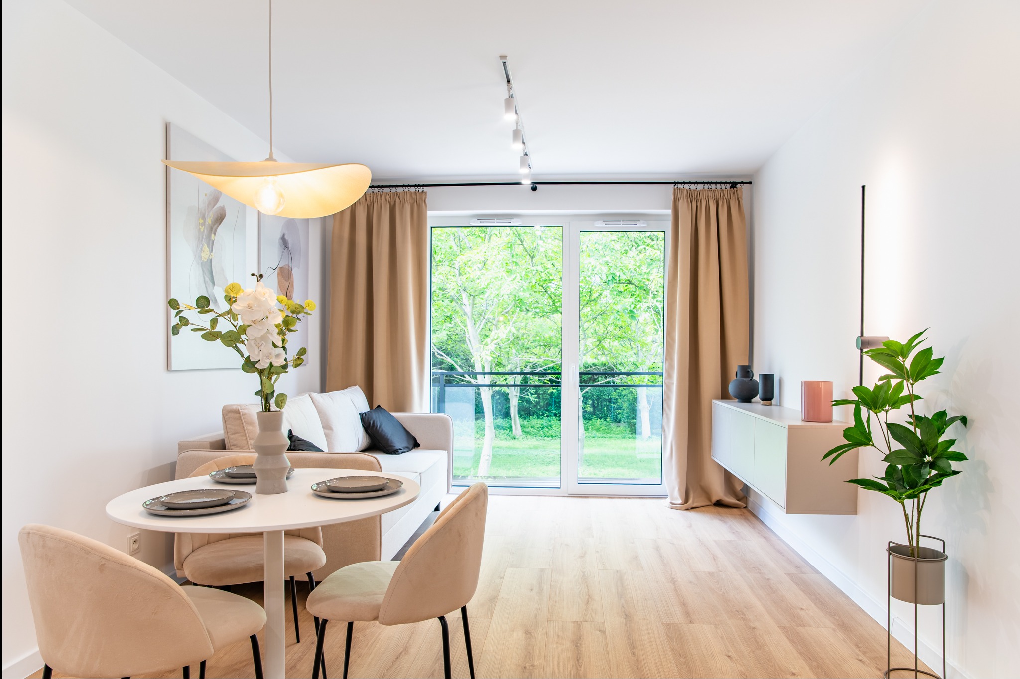 Jasne wnętrze salonu z okrągłym stołem, beżową sofą i dużym oknem balkonowym z widokiem na zieleń. Minimalistyczny design, drewniana podłoga i nowoczesne oświetlenie.