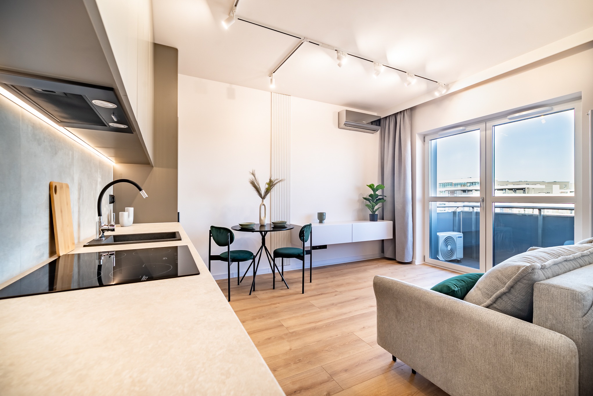 Jasne wnętrze mieszkania z aneksem kuchennym, okrągłym stołem z krzesłami i szarą sofą. Widok na balkon przez duże okno. Minimalistyczny design, oświetlenie punktowe.