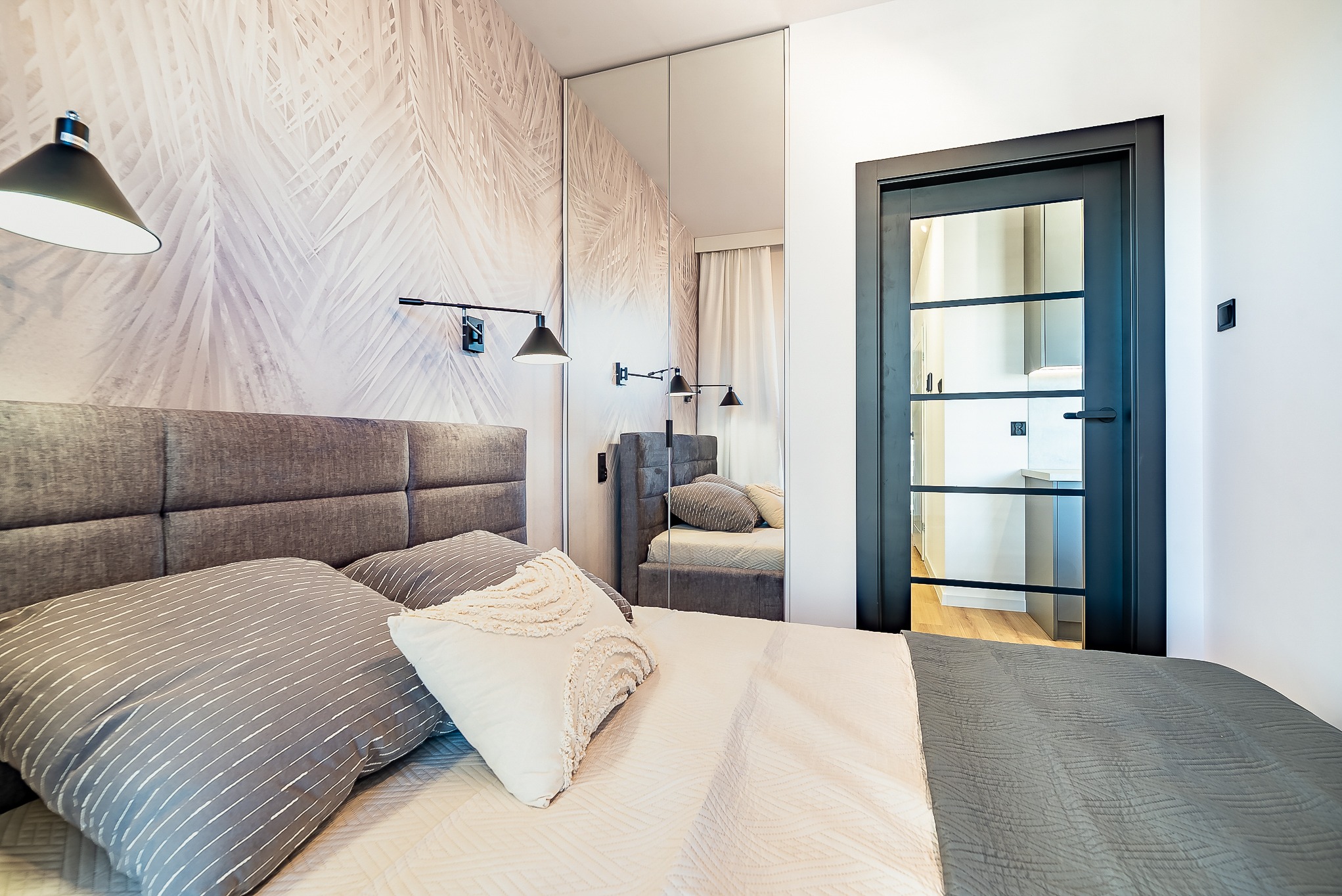 Nowoczesna sypialnia z łóżkiem w odcieniach szarości, tapetą w liście palmowe i czarnymi lampami. Lustrzana szafa wnękowa i czarne drzwi z przeszkleniami.